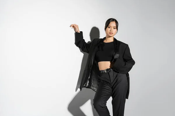 Fashionable asian woman in black crop top and pantsuit looking at camera on grey background with shadow — Stock Photo
