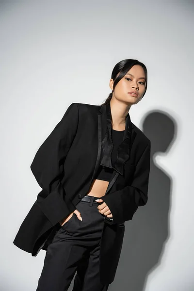 Brunette asian woman in black fashionable suit posing with hand in pocket on grey background with shadow — Stock Photo