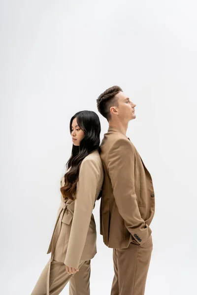 Side view of interracial couple in beige suits standing back to back isolated on grey — Stockfoto