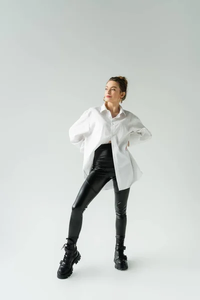 Full length of woman in white oversize shirt and black tight pants posing with hands on waist on grey background — Photo de stock