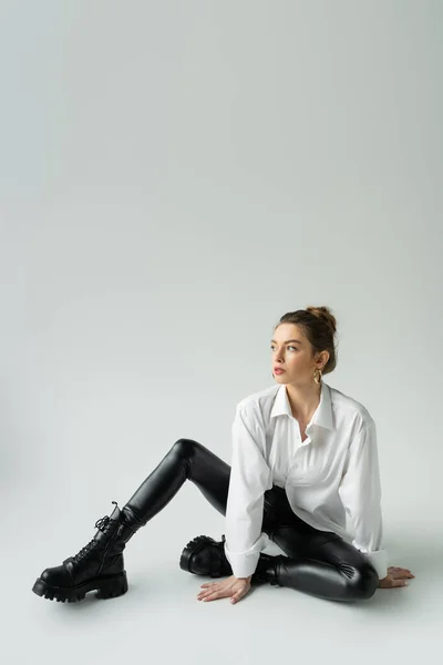 Full length of trendy woman in white shirt and black tight pants sitting and looking away on grey background — Photo de stock