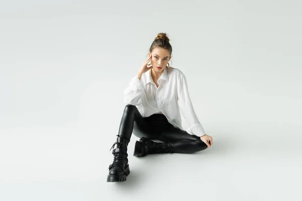 Full length of young model in black latex pants and rough boots sitting on grey background — Stock Photo