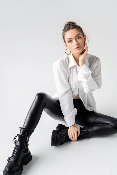 Full length of young woman in white shirt and black latex pants with rough boots sitting on grey background — Photo de stock