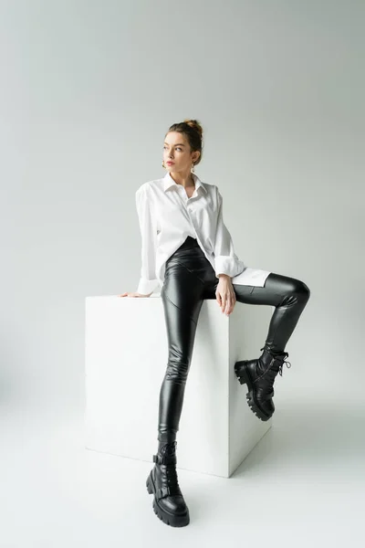 Full length of model in white oversize shirt and black pants with rough boots sitting on white cube on grey background — Stock Photo