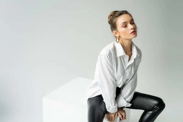 Young sensual woman with closed eyes wearing white shirt and black latex pants and sitting on cube isolated on grey — Stock Photo