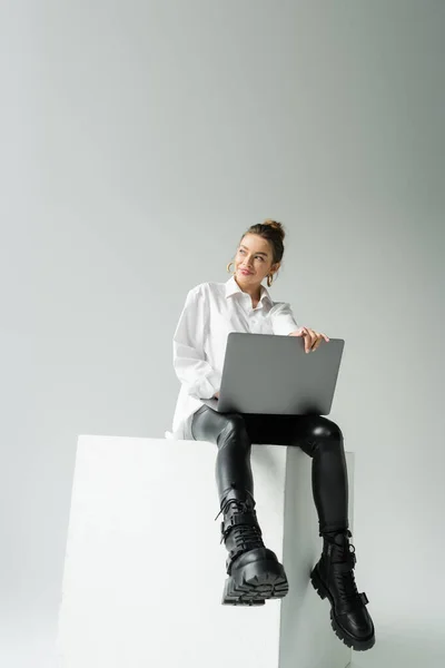 Volle Länge der glücklichen Frau in schwarzen Latexhosen und Lederstiefeln sitzt auf einem weißen Würfel mit Laptop auf grauem Hintergrund — Stockfoto