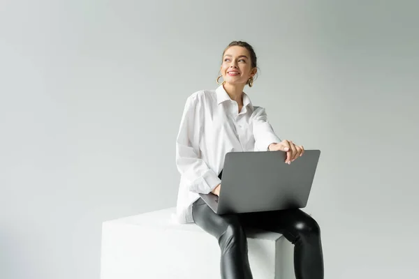 Весела жінка в модному вбранні сидить на білому кубі з ноутбуком і дивиться ізольовано на сірому — стокове фото