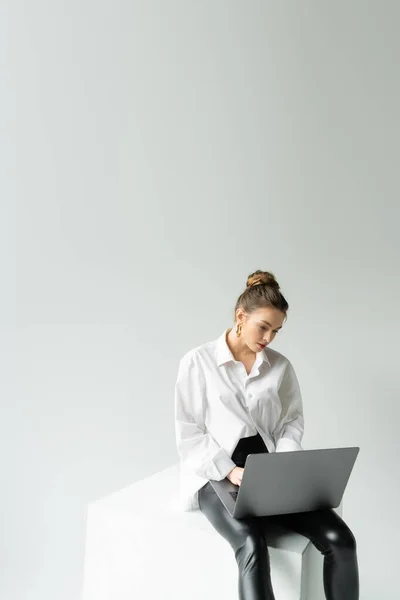 Young woman in oversize shirt and black tight pants working on laptop while sitting on cube on grey background — Stock Photo