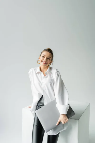 Joyful woman in hoop earrings and oversize shirt holding laptop and looking at camera near white cube on grey background — Stockfoto