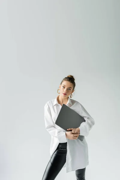 Trendy woman in white shirt and hoop earrings posing with laptop and looking at camera isolated on grey — Stock Photo
