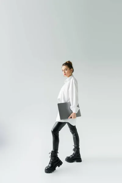 Повна довжина стильної жінки в чорних латексних штанях і грубих чоботях позує з ноутбуком на сірому фоні — стокове фото