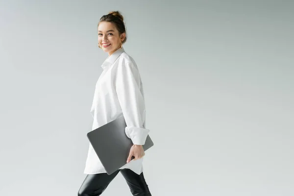 Усміхнена жінка в білій сорочці дивиться на камеру під час ходьби з ноутбуком ізольована на сірому — стокове фото
