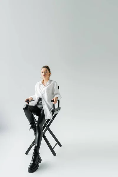 Volle Länge der jungen Frau in modischem Outfit sitzt auf einem Stuhl und schaut in die Kamera auf grauem Hintergrund — Stockfoto