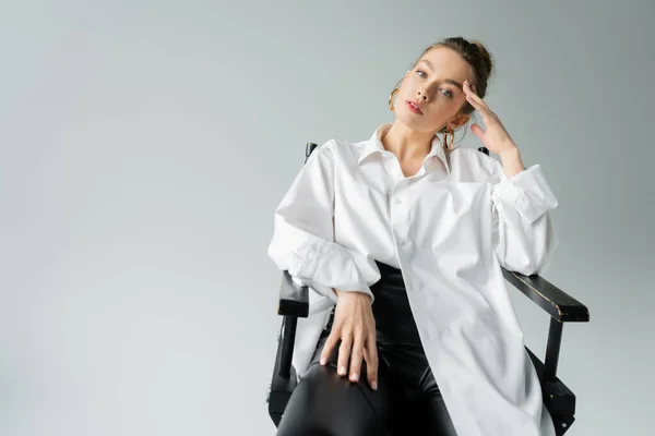 Pretty woman in white oversize shirt and black tight pants posing on chair with hand near head isolated on grey — Stock Photo