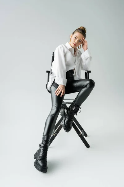 Volle Länge der stilvollen Frau in weißem Hemd und schwarzer Hose mit Lederstiefeln sitzt auf Stuhl auf grauem Hintergrund — Stockfoto