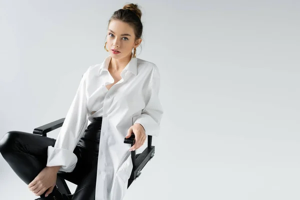 Trendy woman in black tight pants and white oversize shirt sitting on chair and looking at camera isolated on grey — Stock Photo