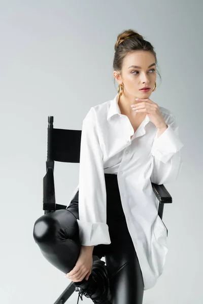 Fashionable woman in white shirt and black latex pants sitting on chair and looking away isolated on grey — Stock Photo