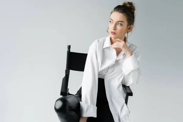 Jeune femme en chemise oversize et pantalon noir serré tenant la main près du menton tout en étant assis sur une chaise isolée sur gris — Photo de stock