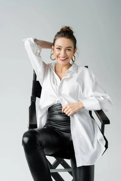 Cheerful woman in white shirt and black latex pants smiling at camera while posing on chair isolated on grey — Stock Photo