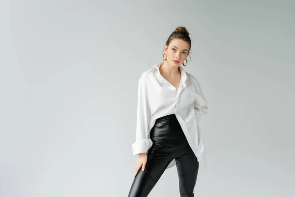 Stylish woman in black tight pants and white oversize shirt posing and looking at camera isolated on grey — Stock Photo