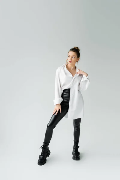 Full length of trendy woman in oversize shirt and tight pants with rough boots looking away on grey background — Stock Photo
