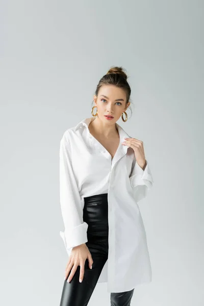 Fashionable woman in white oversize shirt and golden earrings looking at camera isolated on grey — Stockfoto