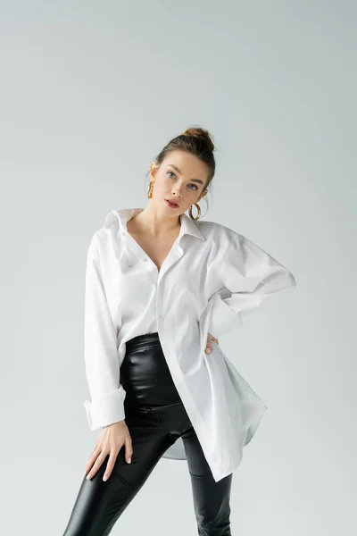 Donna alla moda in pantaloni stretti neri e camicia oversize bianca in posa con mano sul fianco isolata su grigio — Foto stock