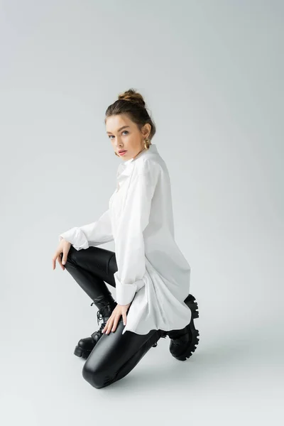 Full length of young woman in white shirt and black rough boots looking at camera while posing on grey background — Stockfoto