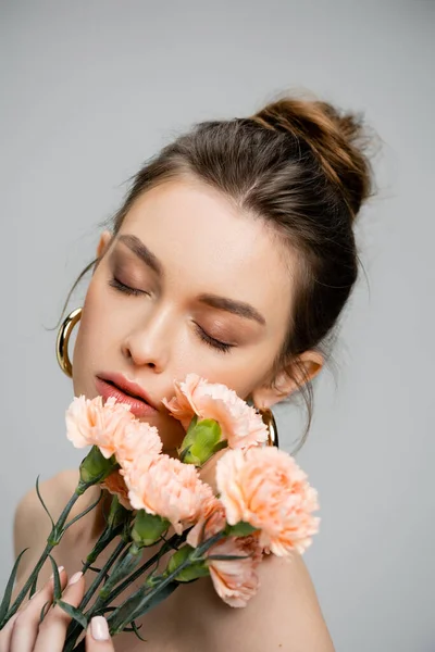 Retrato de mujer joven sensual posando con los ojos cerrados cerca de ramo de claveles de melocotón aislados en gris - foto de stock