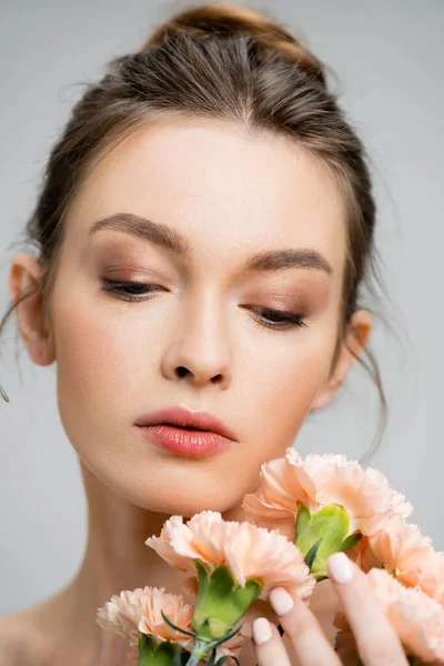 Портрет очаровательной женщины с натуральным макияжем, смотрящей на букет персиковых гвоздик, изолированных на сером — стоковое фото