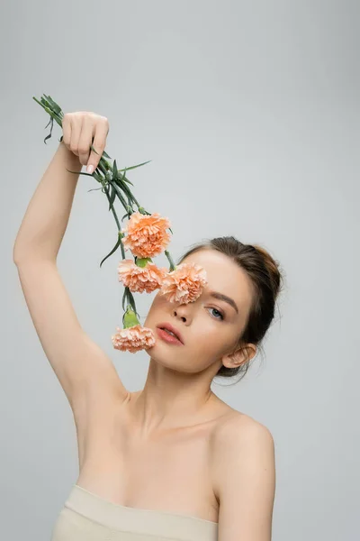 Young woman with naked shoulders obscuring face with pink carnations isolated on grey — Stock Photo