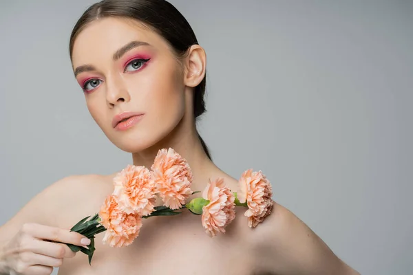 Young woman with makeup and bare shoulders posing with fresh flowers and looking at camera isolated on grey — Stock Photo
