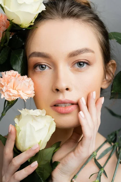 Портрет красивої жінки з ідеальною шкірою, що торкається обличчя біля троянд і гвоздик ізольовані на сірому — стокове фото
