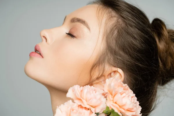 Vista lateral de una joven mujer de pelo rubio posando cerca de claveles borrosos aislados en gris - foto de stock