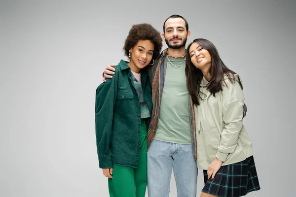 Stylish bearded man hugging young interracial women and smiling at camera isolated on grey — Stock Photo