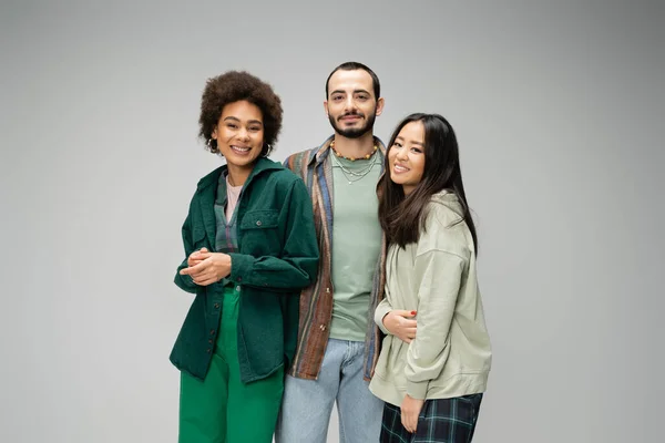 Junge und glückliche multikulturelle Freunde in stylischer Freizeitkleidung schauen vereinzelt in die Kamera auf grau — Stockfoto
