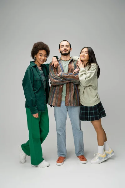 Full length of pleased bearded man posing with crossed arms near young multiethnic women on grey background — Stock Photo