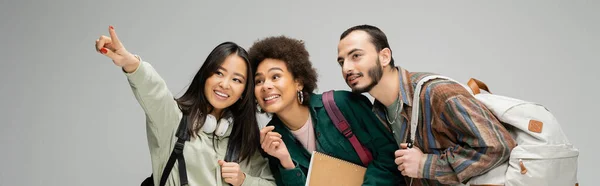 Smiling asian woman looking away and pointing with finger near multiethnic students isolated on grey, banner — Stockfoto