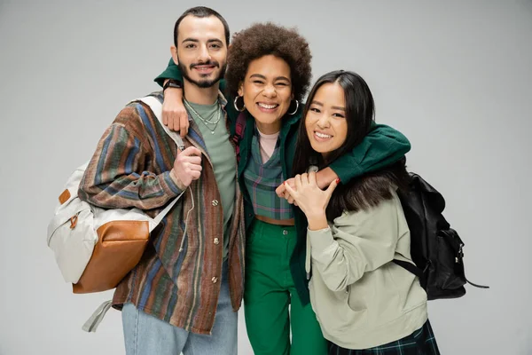 Mulher americana africana animado abraçando estudantes multiétnicos elegantes com mochilas isoladas em cinza — Fotografia de Stock