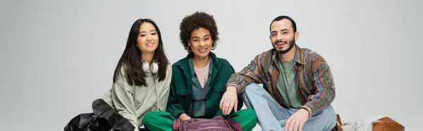 Happy multicultural students in trendy clothes sitting and looking at camera isolated on grey, banner — Foto stock