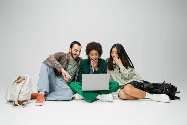 Full length of amazed multicultural friends looking at laptop while sitting with crossed legs on grey background — Photo de stock