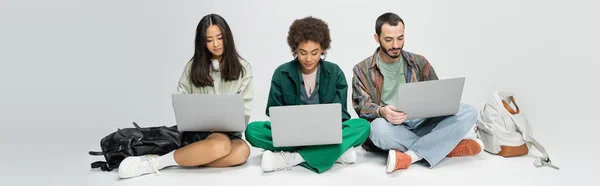 Volle Länge der stilvollen multiethnischen Studenten mit Laptops, während sie in der Nähe von Rucksäcken auf grauem Hintergrund sitzen, Banner — Stockfoto