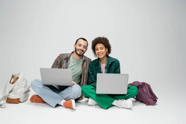 Trendige interrassische Studenten lächeln in die Kamera, während sie mit Laptops in der Nähe von Rucksäcken auf grauem Hintergrund sitzen — Stockfoto