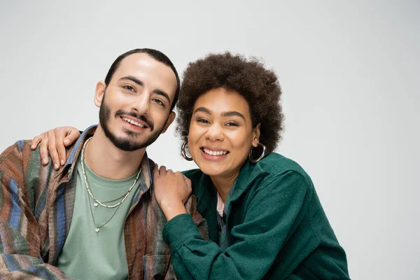 Pretty african american woman smiling at camera and hugging shoulders of bearded man isolated on grey — Stock Photo