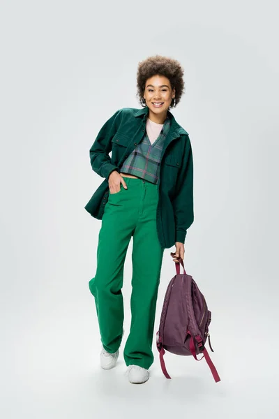 Full length of stylish african american woman holding backpack while posing with hand in pocket of green pants on grey background — Stock Photo