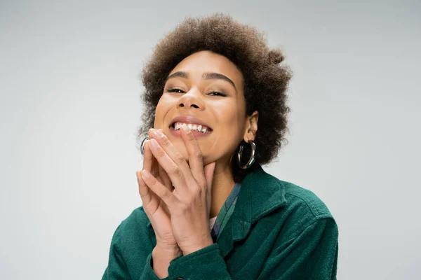 Portrait of excited african american woman with curly brunette hair holding hands near face and looking at camera isolated on grey — Stock Photo
