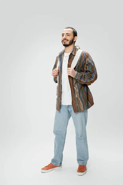 Full length of smiling man in blue jeans and colorful shirt standing with backpack on grey background — Stock Photo