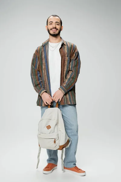 Pleine longueur de heureux étudiant barbu en chemise colorée et jean bleu debout avec sac à dos sur fond gris — Photo de stock