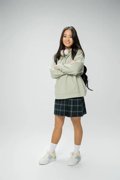Full length of pretty asian student in checkered skirt posing with crossed arms on grey background — Stock Photo