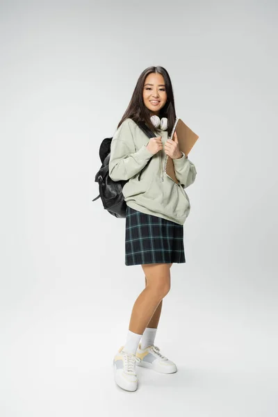 Piena lunghezza di felice donna asiatica in abito elegante in piedi con zaino e notebook su sfondo grigio — Foto stock